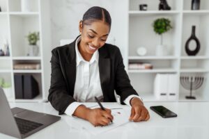 Happy black female business analyst writing on papers, checking data, charts and graphs, sitting at